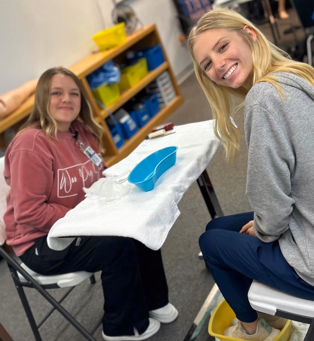 Two smiling PAL CNA students practicing personal care skills in the training lab